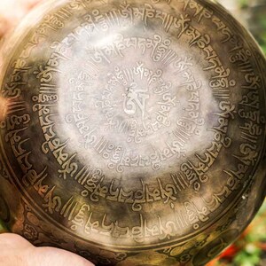 May include: A hand holds a large, round, metal singing bowl with intricate engraved designs and Sanskrit script. The bowl is dark brown with gold accents.