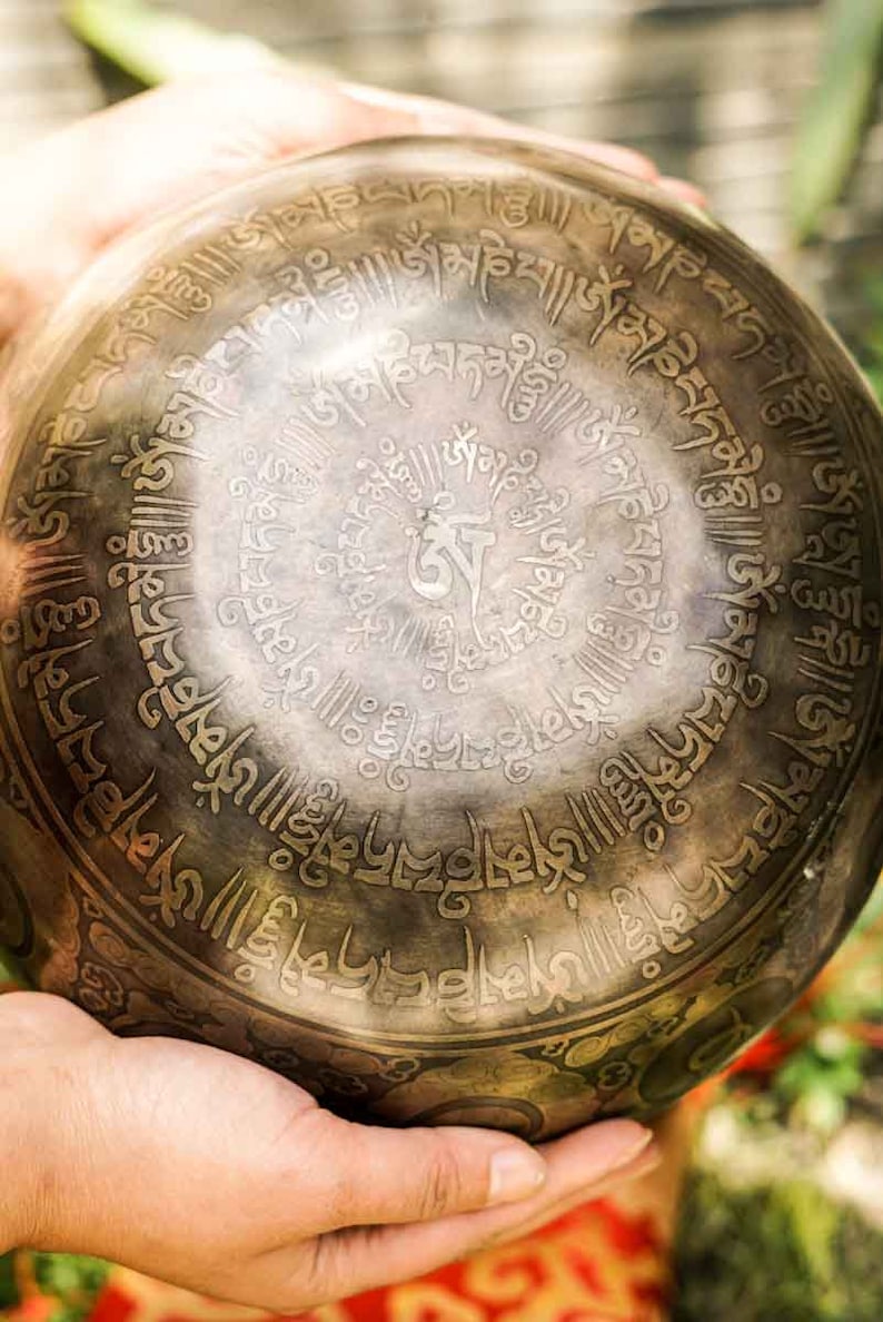 May include: A hand holds a large, round, metal singing bowl with intricate engraved designs and Sanskrit script. The bowl is dark brown with gold accents.