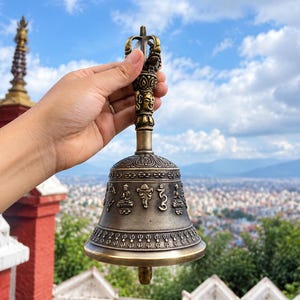 Könnte beinhalten: Eine rituelle Glocke aus bronzefarbenem Metall mit detaillierten Prägungen von sitzenden Figuren und Symbolen am bauchigen Körper. Der Griff ist mit einem dekorativen Vajra-Endstück versehen und zeigt ein skulptiertes Gesicht am Schaft.