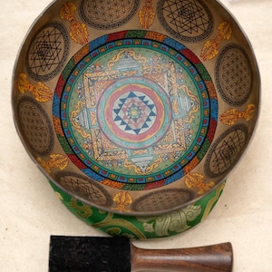 May include: A detailed singing bowl featuring geometric and mandala patterns in brown, blue, green, and red. The bowl sits on a green cushion, with a wooden mallet. Prayer flags are visible in the background.