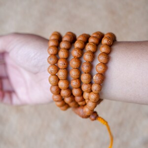 May include: A brown wooden mala bead necklace with a yellow tassel. The necklace is wrapped around a person's wrist.