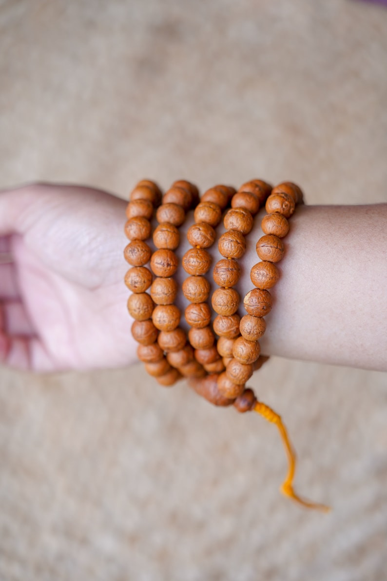 May include: A brown wooden mala bead necklace with a yellow tassel. The necklace is wrapped around a person's wrist.