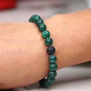 May include: A green malachite beaded bracelet with a black lava bead. The bracelet is worn on a person's wrist.