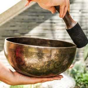 May include: A person holds a large, ornate, brass singing bowl with a wooden striker. The bowl has intricate designs and a circular symbol in the center. The bowl is being struck with the striker, creating a sound.