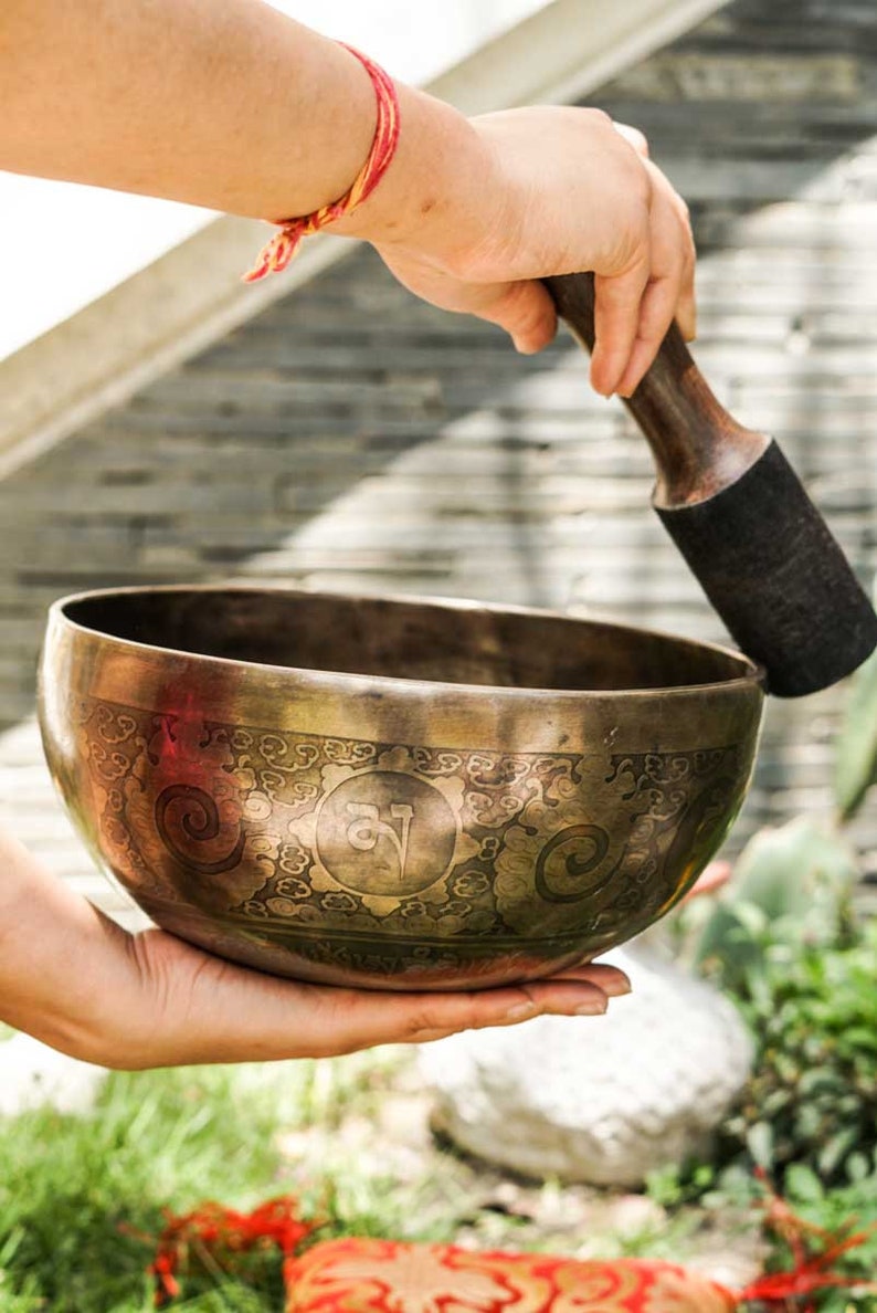 May include: A person holds a large, ornate, brass singing bowl with a wooden striker. The bowl has intricate designs and a circular symbol in the center. The bowl is being struck with the striker, creating a sound.