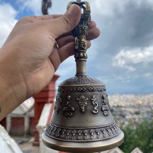 May include: Bronze-colored bell with intricate embossed designs and a detailed handle featuring a face and crown. The bell's surface displays religious symbols and patterns. The bell is held against a backdrop of a cloudy sky and a distant cityscape.