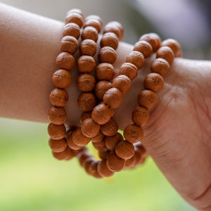 May include: A close-up of a brown wooden beaded bracelet worn on a person's wrist. The bracelet is made of multiple strands of beads, creating a layered effect.