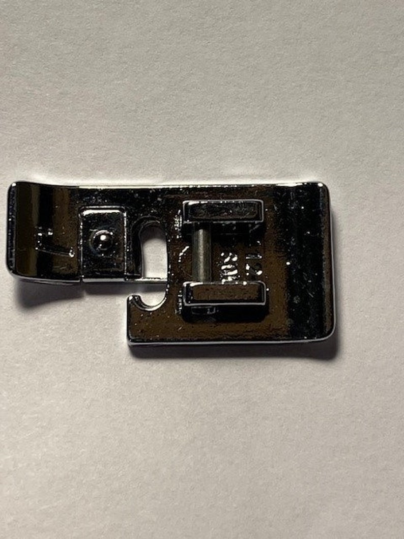 K&ouml;nnte beinhalten: Ein silberner N&auml;hmaschinenfu&szlig; mit einer Metallstange und einem kleinen Loch in der Mitte.