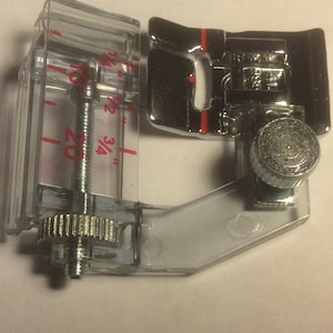 May include: A clear plastic sewing foot with a metal screw and a silver knob. The foot has a ruler on the side with markings in inches and fractions of an inch. The foot is designed to be used with a sewing machine.