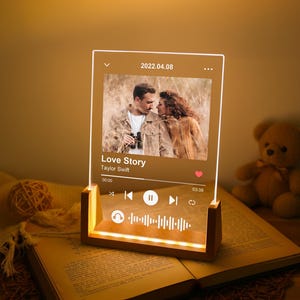 Può includere: Una luce notturna in acrilico trasparente con base in legno. Mostra una foto di una coppia e il testo "Love Story" di Taylor Swift. È visibile anche la data "2022.04.08". La luce è su un libro aperto.