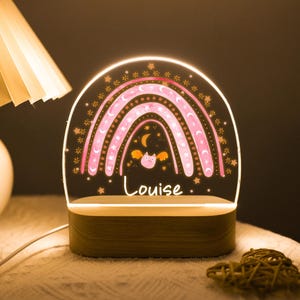 May include: A decorative night light featuring a pink rainbow design with stars and a bat. The name "Louise" is written in cursive. The light sits on a wooden base and is illuminated.