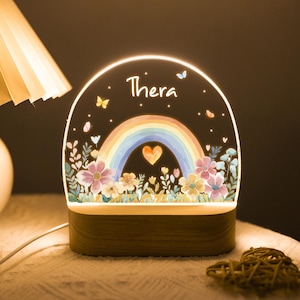 Op de afbeelding: Decoratief nachtlampje met een regenboogontwerp, een hart en bloemen. De naam "Thera" staat bovenaan. Het staat op een houten voet en straalt een warme gloed uit.