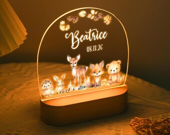 May include: A personalized night light with a wooden base and a clear acrylic top. The acrylic features the name "Beatrice" and a date, along with illustrations of woodland animals. The light emits a warm glow.