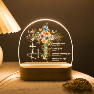 Puede incluir: Una luz nocturna personalizada con un diseño de cruz floral. La luz presenta el nombre "Amelia" y la fecha "12.09.2025". La cruz está adornada con flores coloridas e incluye versículos bíblicos. La base es de madera.