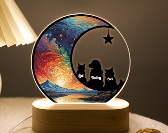Pode incluir: Uma luz noturna de madeira com um design de lua crescente. A lua está cheia de uma cena de galáxia colorida e apresenta três silhuetas de animais de estimação: um cão chamado Bailey e dois gatos chamados Lexi e Coco.