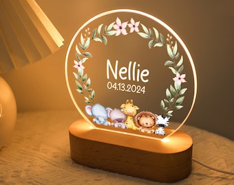 May include: A white acrylic night light with a floral wreath design and the name "Nellie" and the date "04.13.2024" printed on it. The light features a wooden base and four small animal illustrations: an elephant, a hippopotamus, a giraffe, and a zebra.