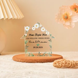 May include: A clear acrylic house-shaped sign with the text "Home Begins Here," address, names, and a date. The sign is decorated with white daisies and green leaves. It is displayed on a round, brown cork base.