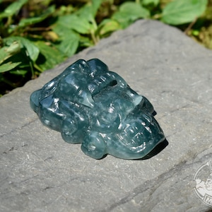 May include: A translucent, dark teal-coloured stone carving shaped like a stylised skull with prominent teeth and intricate facial features. The object has a smooth, polished surface and is positioned on a flat, grey, textured stone surface.