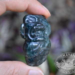 May include: A hand holds a carved stone figurine of a Mayan figure. The figurine is dark teal and has detailed facial features. The background is blurred with green and purple hues. The Mayan Mountain logo is visible.