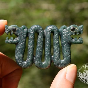 May include: A dark green carved stone pendant featuring two serpent-like creatures with open mouths and textured scales, connected in a continuous wavy pattern. The piece includes a logo in the corner with the text MAYAN MOUNTAIN.
