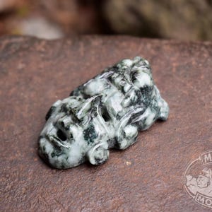 May include: A carved jade figurine in shades of green and white, depicting a stylized animal or mythical creature. The sculpture is detailed with visible facial features and sits on a brown surface. The Mayan Mountain logo is visible.