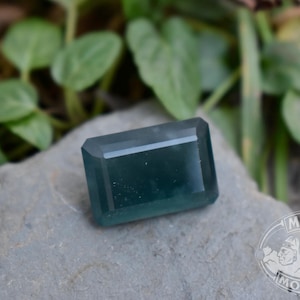 Puede incluir: Una gema rectangular de color verde azulado con un corte facetado. La gema se muestra sobre una superficie de piedra gris, con follaje verde en el fondo. La gema tiene un aspecto pulido.
