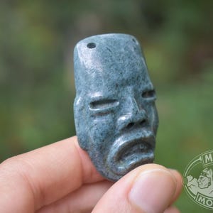 Op de afbeelding: Een gebeeldhouwde, blauwgroene stenen hanger die een gestileerd menselijk gezicht afbeeldt met gesloten ogen en een neerhangende mond. De hanger heeft twee gaten aan de bovenkant en is waarschijnlijk een Maya-artefact.