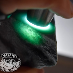 May include: A close-up of a dark gray stone illuminated by a bright green light. The light source is a ring shape, highlighting the stone's texture and color. The Mayan Mountain logo is visible.