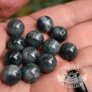 Puede incluir: Una colección de cuentas de piedra redondas y pulidas en tonos gris oscuro y negro con veteado natural visible y pequeños orificios de perforación centrales. Las cuentas tienen una textura superficial lisa y reflectante. Un logotipo con el texto MAYAN MOUNTAIN es visible en la esquina inferior derecha.