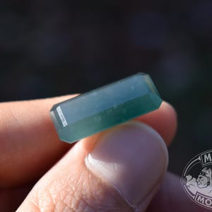 Op de afbeelding: Een rechthoekige, facetgeslepen, teal-kleurige edelsteen. De edelsteen wordt in een hand gehouden, met een vage achtergrond. Het Mayan Mountain-logo is zichtbaar in de rechter benedenhoek.