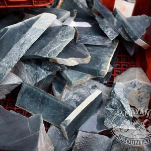 May include: A collection of raw, irregularly shaped jade stones in shades of green and gray. The stones are piled inside a red plastic crate, with some pieces displaying a polished surface. The image includes the "Mayan Mountain" logo.