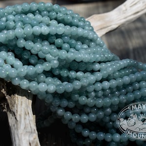 May include: Strands of round, pale blue-green beads. The beads are draped over a piece of driftwood. The logo for Mayan Mountain is visible in the lower right corner. The beads are a soft, muted color.