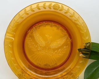 May include: Amber glass ashtray with an embossed design of an eagle with outstretched wings, surrounded by stars and a wreath. The ashtray has a leaf and flower design around the rim.