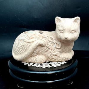 May include: A beige ceramic cat figurine in a reclining position, featuring embossed floral and ribbon details along its back and side. The cat has two circular openings on its upper back and is displayed on a round, black, elevated base.