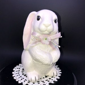 May include: A white ceramic bunny figurine with pink ears and a pink bow with a small pink flower. The bunny is sitting with its paws in front of it.