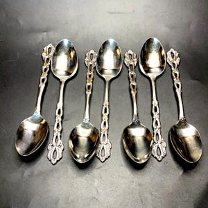 May include: Six silver spoons with ornate handles and oval bowls are arranged on a dark surface. The handles feature a decorative openwork design with a floral motif at the top. The spoons are reflective, showing the surrounding environment.