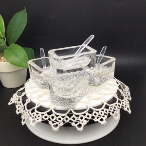 May include: Four clear glass condiment dishes with matching clear plastic spoons. The dishes are arranged on a white doily on a black and white pedestal.