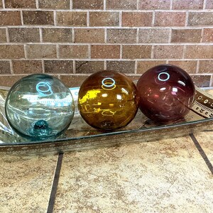 May include: A rectangular glass tray with a decorative border holds three spherical glass floats. The floats are translucent and colored in shades of light blue, amber, and deep purple, each featuring a smooth, reflective surface and circular highlights.