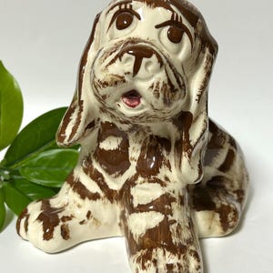 May include: A ceramic figurine of a brown and white puppy with large eyes and a surprised expression. The puppy is sitting with its legs crossed and its ears perked up.