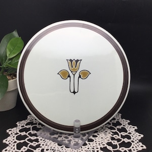 May include: A white ceramic trivet with a brown rim and a stylized flower design in brown and gold.