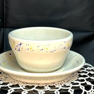 May include: A white ceramic bowl and saucer set with a colorful splatter design in yellow, blue, and red.