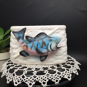 May include: A white ceramic planter shaped like a basket with a blue, pink, and black fish jumping out of it. The fish is painted with realistic details and has a fishing rod coming out of its mouth.
