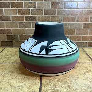 May include: A wide-mouthed ceramic vase featuring a matte black neck and a rounded body. The upper section displays a white band with black geometric shapes, while the lower portion transitions through horizontal gradients of green, purple, and white.