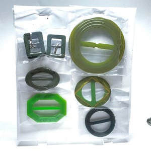 May include: A collection of vintage belt buckles in various shapes and shades of green, including square, oval, and circular designs. The buckles are made of plastic and are displayed in individual plastic sleeves.