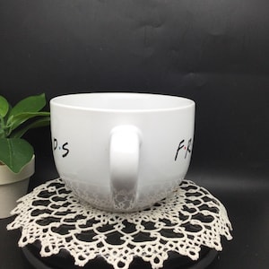 Puede incluir: Taza de cer&aacute;mica blanca con un dise&ntilde;o negro de "FRIENDS". La taza est&aacute; sobre un encaje de ganchillo blanco.