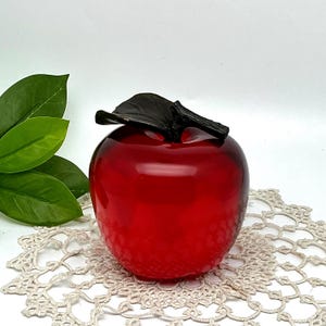 May include: A glossy red apple figurine with a dark leaf and stem. The decorative apple is set on a cream-colored, lace doily. Green leaves are visible in the background. The apple is a classic decorative item.