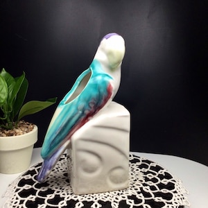 May include: A ceramic parrot planter with a blue, green, white, and purple glaze. The parrot is perched on a white ceramic base with a geometric design.