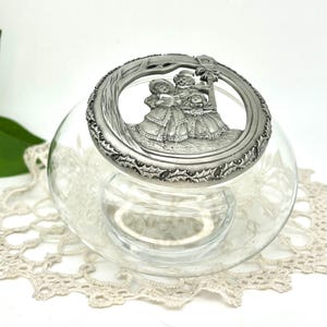 May include: A clear glass jar with a decorative silver lid. The lid features a detailed relief of carolers singing under a tree. The lid is ringed with a holly leaf design. The jar sits on a cream-colored lace doily.