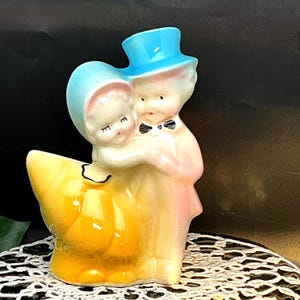 May include: A vintage ceramic figurine of a couple embracing. The man wears a blue top hat and a black bow tie. The woman wears a blue bonnet. The figurine has a yellow base and is painted in pastel colors.