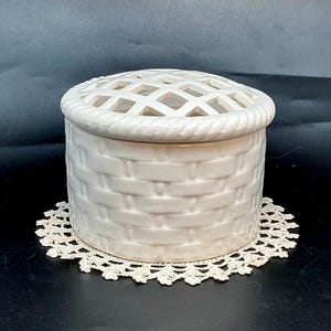 May include: A round, off-white ceramic basket-weave container featuring a removable lid with a lattice-style grid top. The base of the container has a textured, woven-look pattern, and the lid is finished with a decorative twisted rope border.